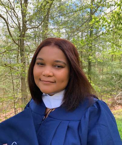 Environmental studies graduate in her gown smiles into the camera with trees in the background, holding her graduation cap. 